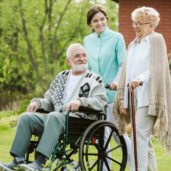 worker and family member caregiving and walking with elderly man in wheelchair - Family Tree Cares